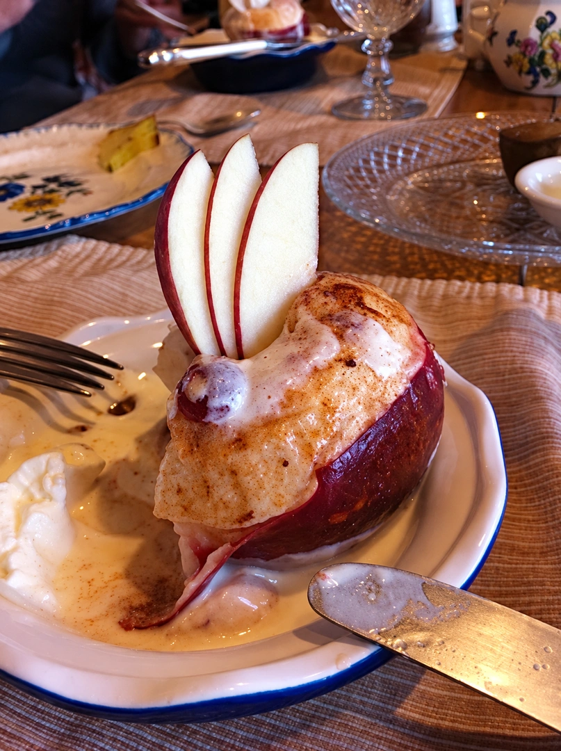 Baked apple dessert topped with cream and fanned apple slices on a plate, a knife at right. Photograph by Louis Zoellar Bickett. Image from the AEQAI archive. (Digitally enhanced. May contain discrepancies from the original artwork.)