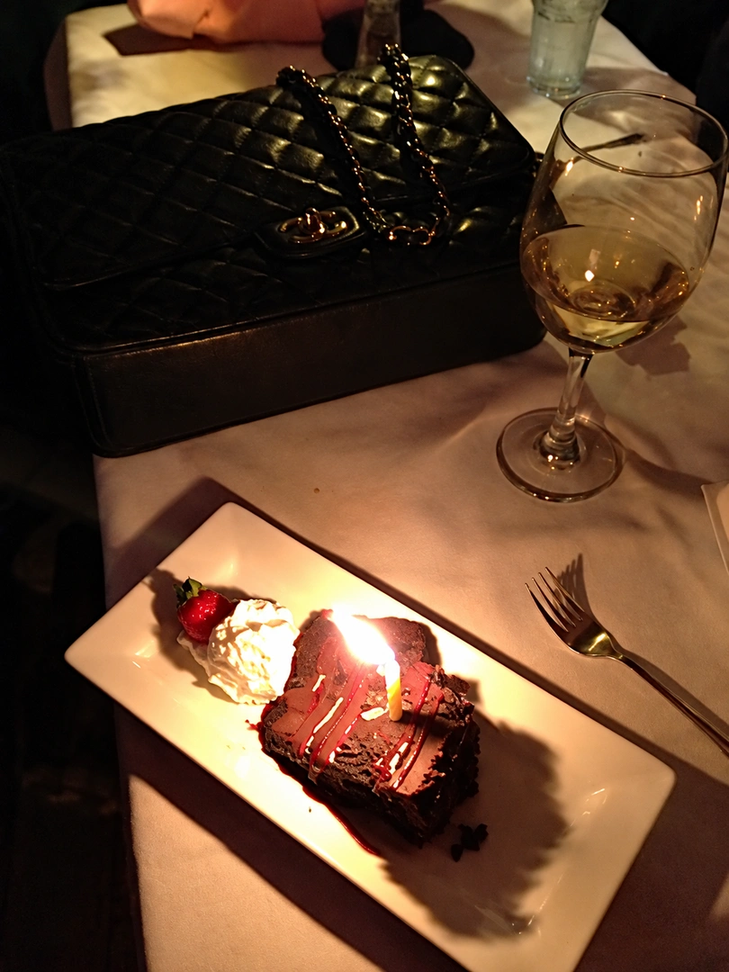 Chocolate dessert with a lit candle on a plate; a wine glass and a handbag on a dimly lit restaurant table. Photograph by Louis Zoellar Bickett. Image from the AEQAI archive. (Digitally enhanced. May contain discrepancies from the original artwork.)