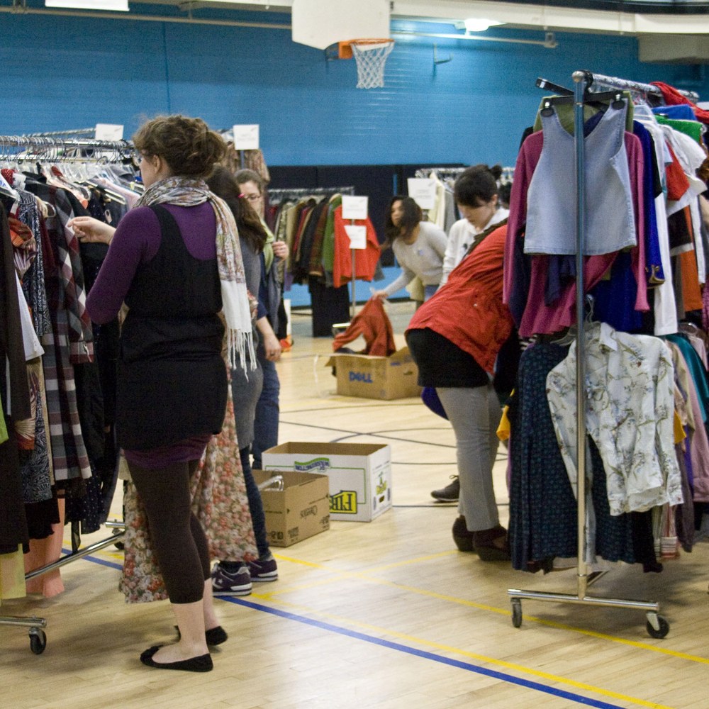 People browsing racks of secondhand clothing at a clothes swap event in a gymnasium.