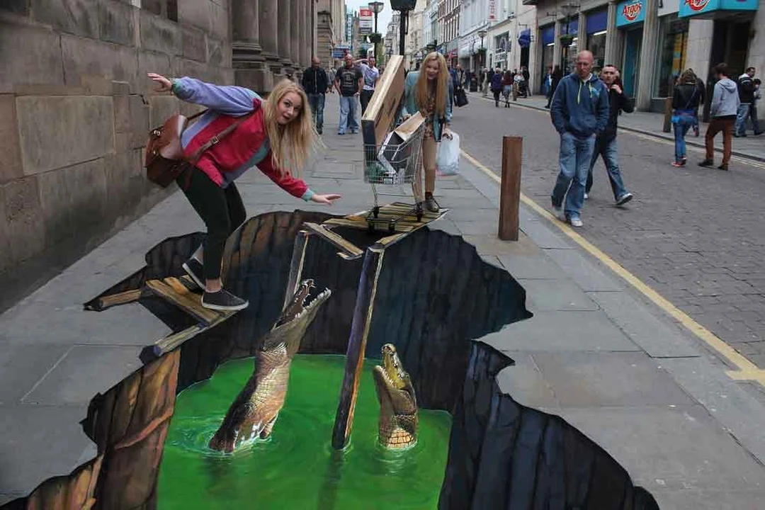 Two women posing beside a 3D street-art painting of two crocodiles rising from a hole painted in a city sidewalk, with onlookers behind.