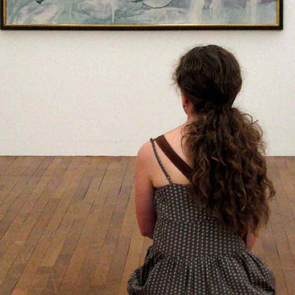 A visitor standing in a gallery, looking slowly at a painting on the wall.