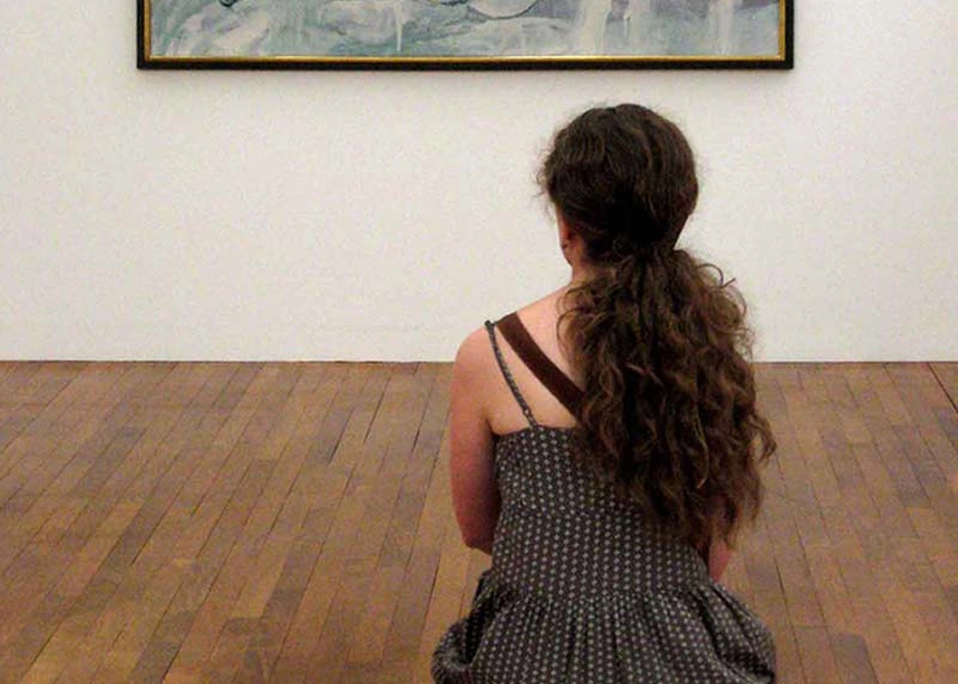 A visitor standing in a gallery, looking slowly at a painting on the wall.