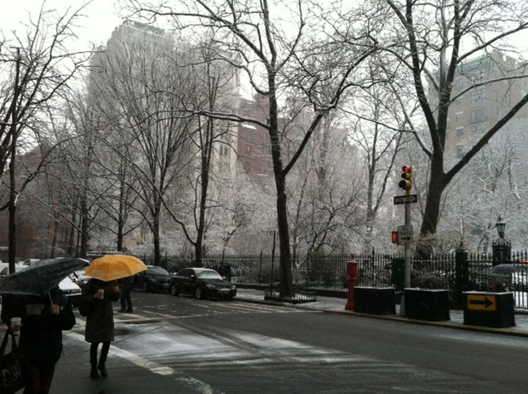 Snow-covered trees line a city street in front of Gramercy Park as pedestrians with umbrellas cross at an intersection