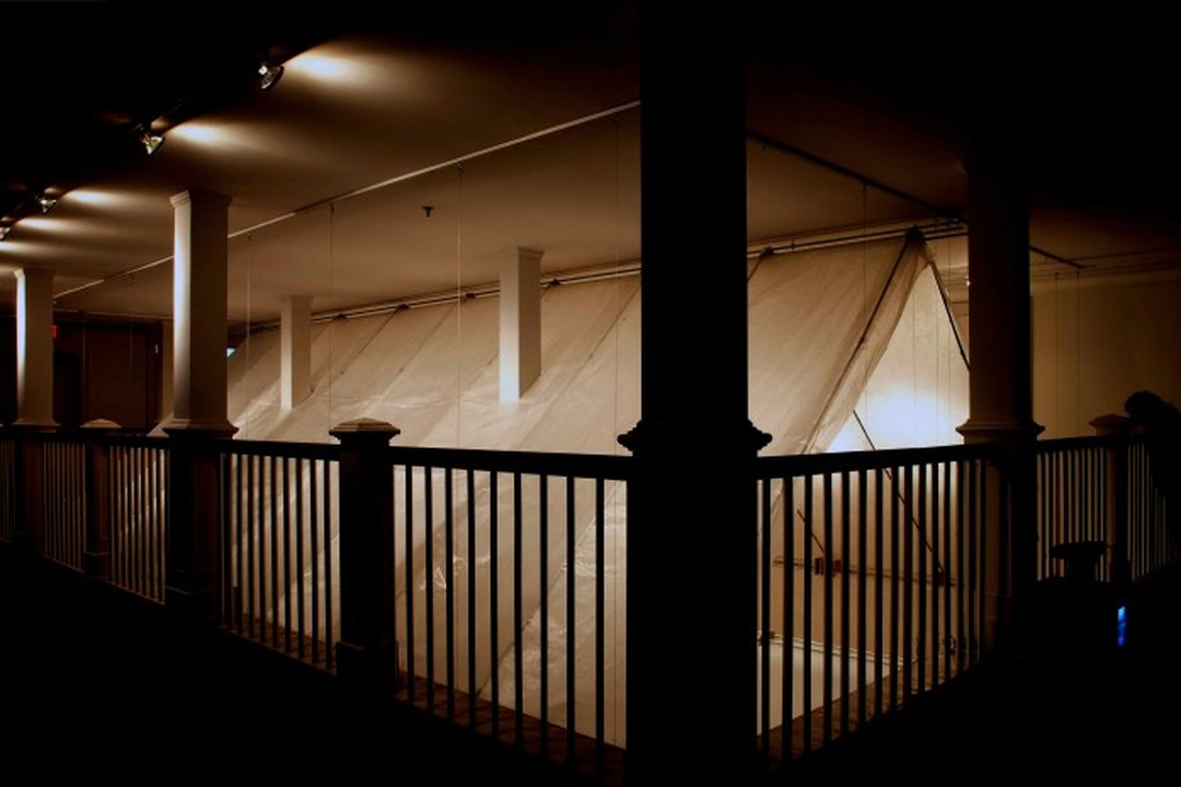 Interior of Michael Casselli's wax paper house installation at Herndon Gallery: white columns and a dark railing in the foreground, translucent glowing walls behind.