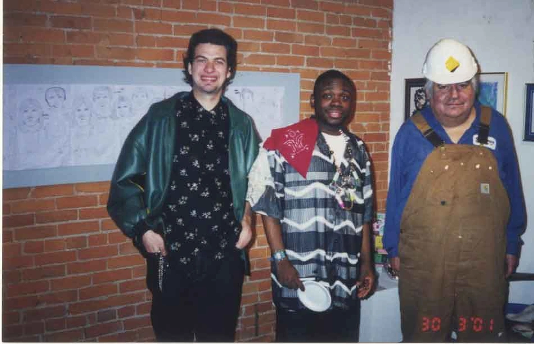 Three men posing in front of a brick wall with a large drawing of faces on the wall behind them; the photograph bears a date stamp reading 30 3 '01 in the lower right corner.