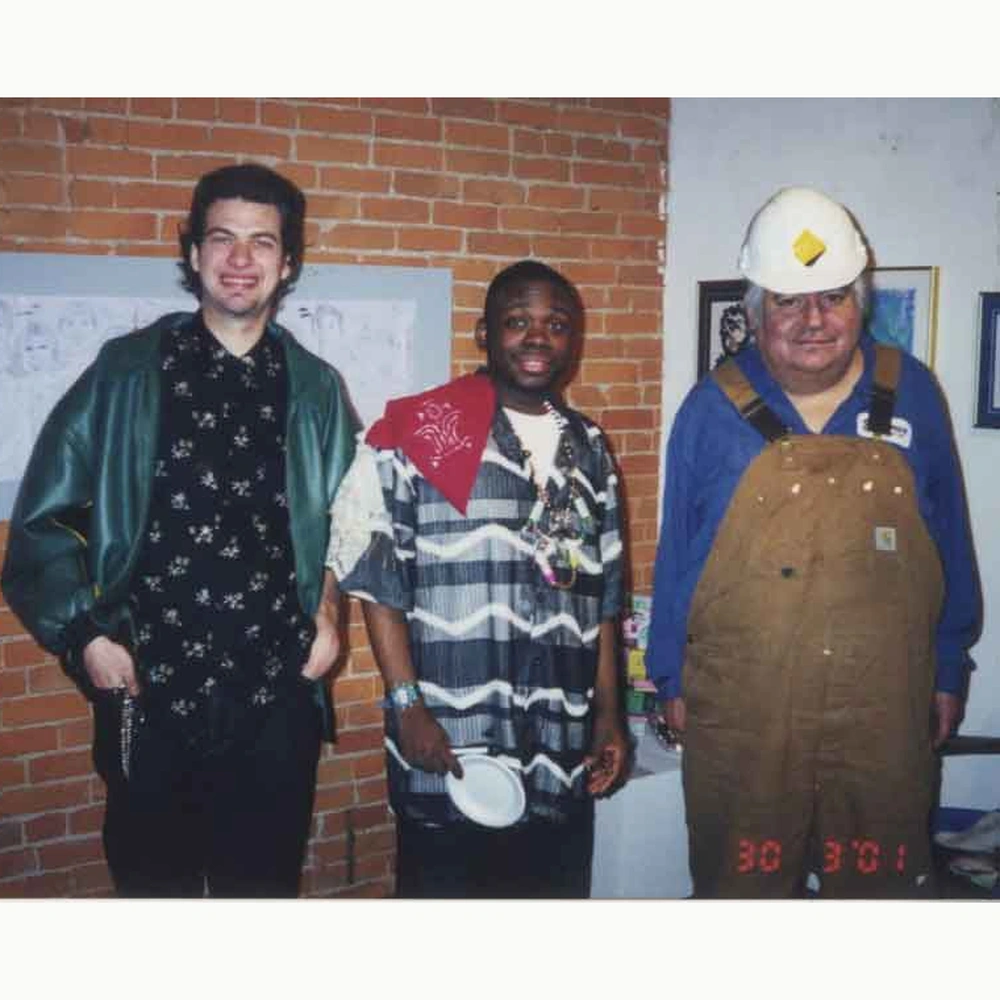 Three men posing in front of a brick wall with a large drawing of faces on the wall behind them; the photograph bears a date stamp reading 30 3 '01 in the lower right corner.