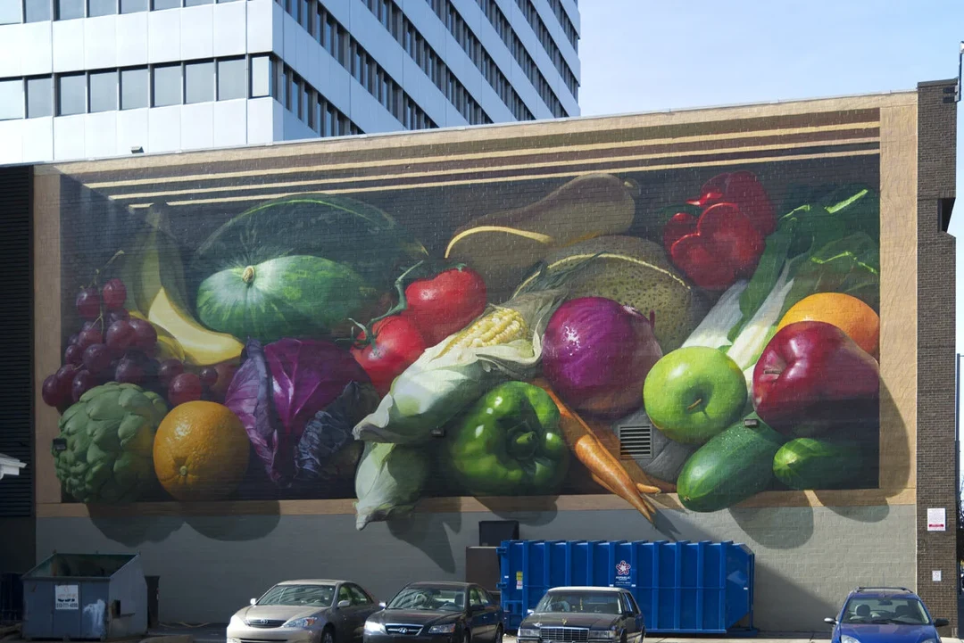 Photorealistic mural of oversized fruits and vegetables in a wooden crate, painted on the side of a building.