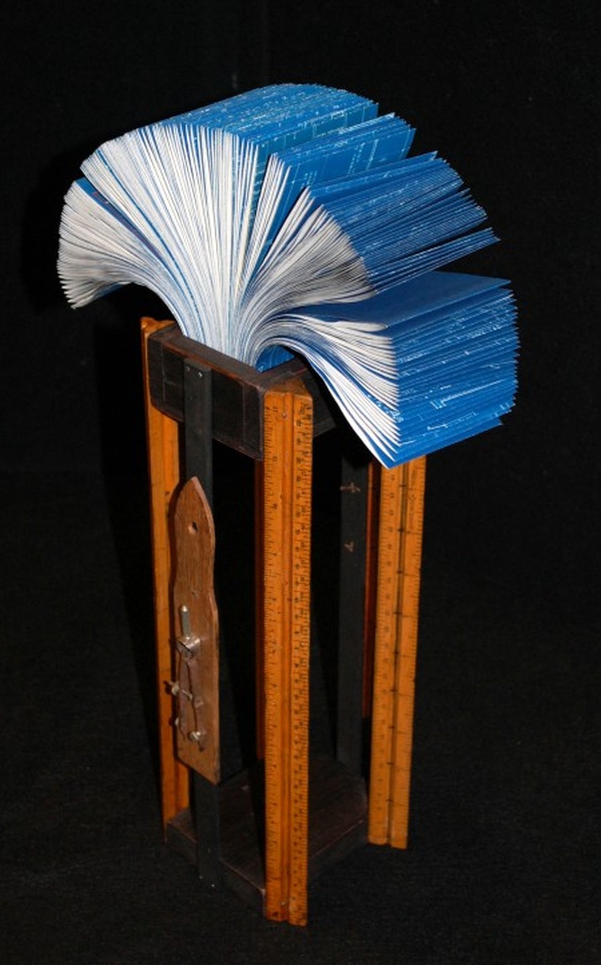 Fan-shaped blue book sculpture mounted on a stand made of wooden rulers, against a black background.