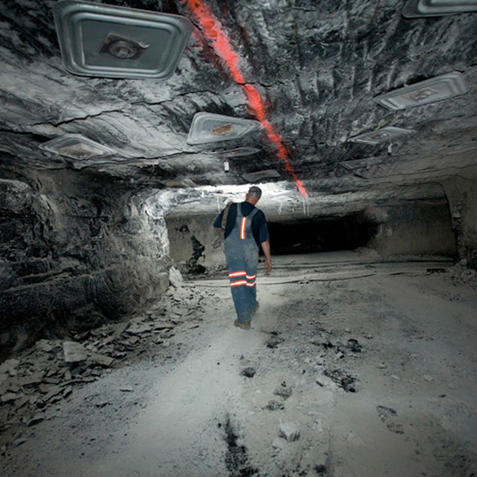 A coal miner wearing overalls with orange reflective stripes walks away from the camera down an underground coal mine tunnel, beneath ceiling lights and a red laser line on the rock roof.