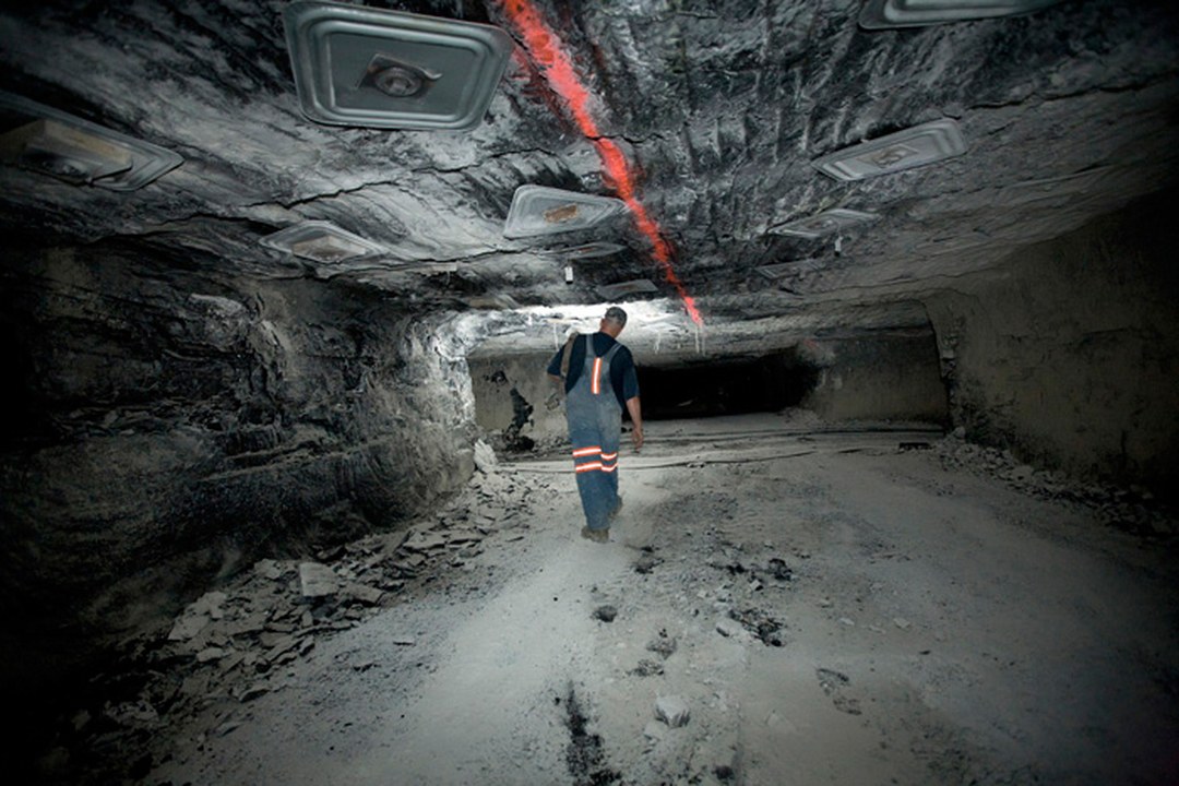 A coal miner wearing overalls with orange reflective stripes walks away from the camera down an underground coal mine tunnel, beneath ceiling lights and a red laser line on the rock roof.