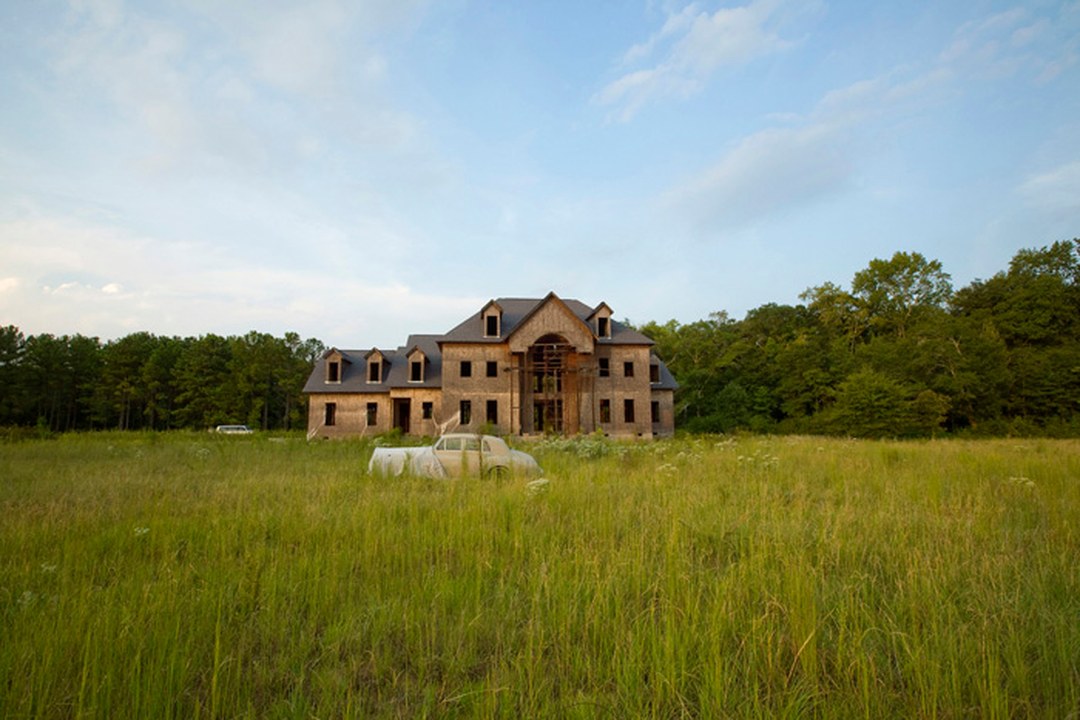 A large unfinished house stands alone in a tall-grass field, with two tarp-covered cars parked in front of it and a line of trees behind, under a pale blue sky.