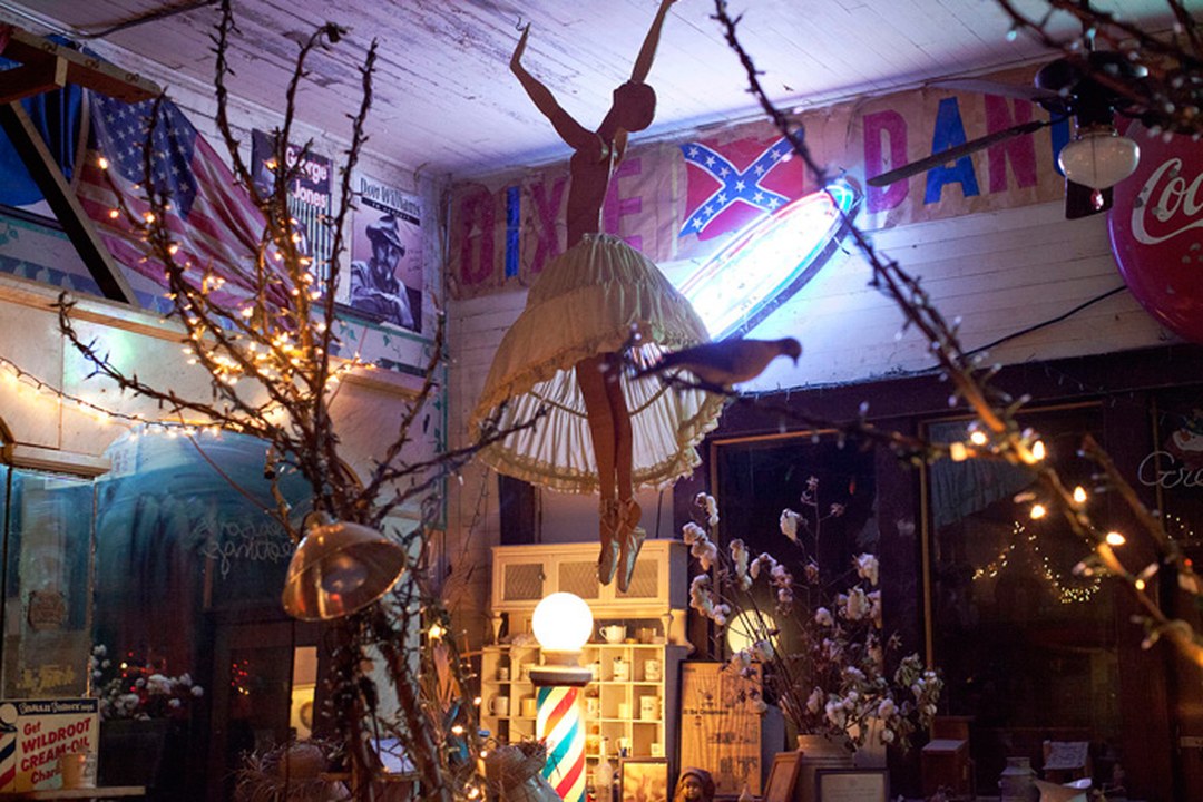 Interior of a Southern antique shop crowded with Americana: a Confederate flag and a “DIXIE” banner hang above a doll figure suspended from the ceiling, surrounded by string lights and vintage signs.