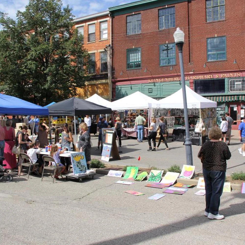 Artist booths and visitors at an outdoor street art fair in Covington, with paintings displayed on the pavement in front of brick buildings.