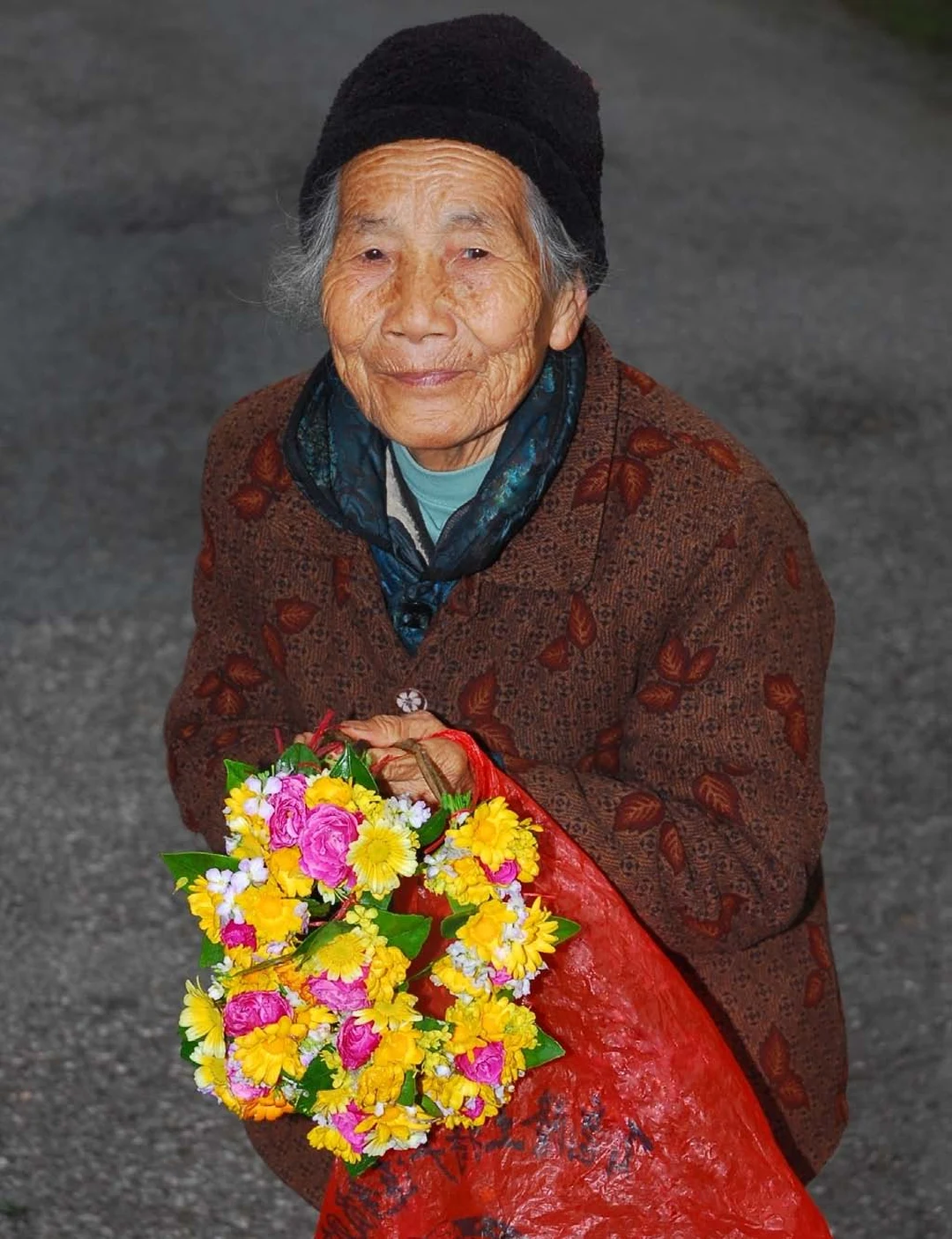 Elderly woman in a dark knit cap and brown coat holding a large bouquet of pink, yellow and white flowers, smiling at the camera.