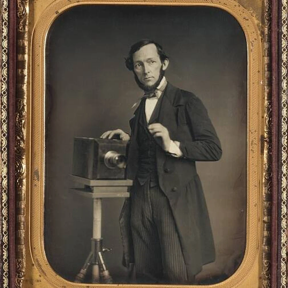 Man in a long jacket standing beside a camera on a tripod, hand raised as if conducting, in a half-plate daguerreotype.