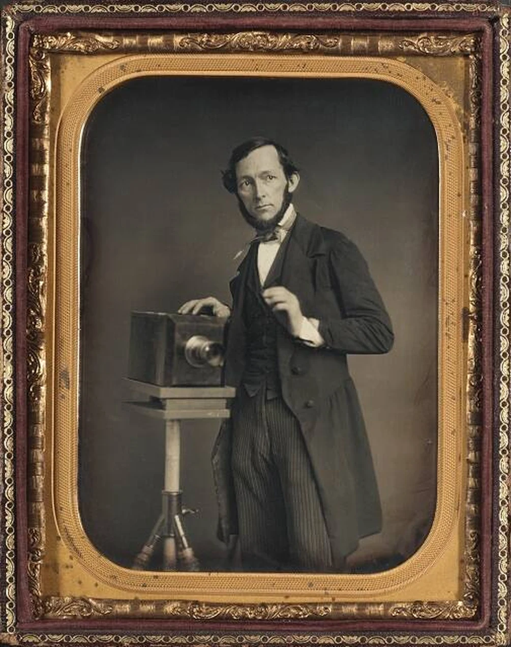 Man in a long jacket standing beside a camera on a tripod, hand raised as if conducting, in a half-plate daguerreotype.