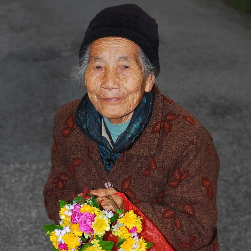 Elderly woman in a dark knit cap and brown coat holding a large bouquet of pink, yellow and white flowers, smiling at the camera.