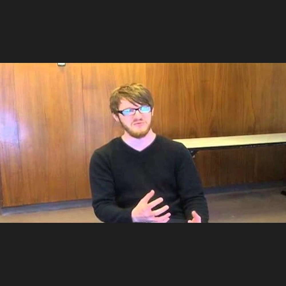 Young man with glasses speaking during a video interview, seated in front of wood paneling.
