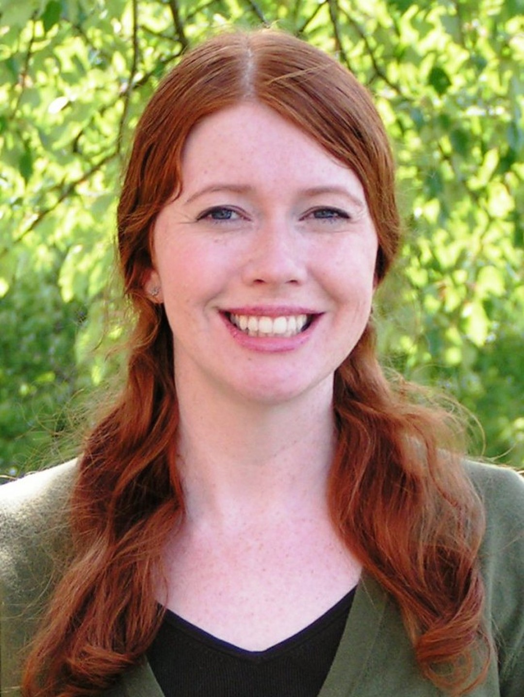 Headshot portrait of Emma Caro, a smiling woman with long auburn hair, photographed outdoors against green foliage.