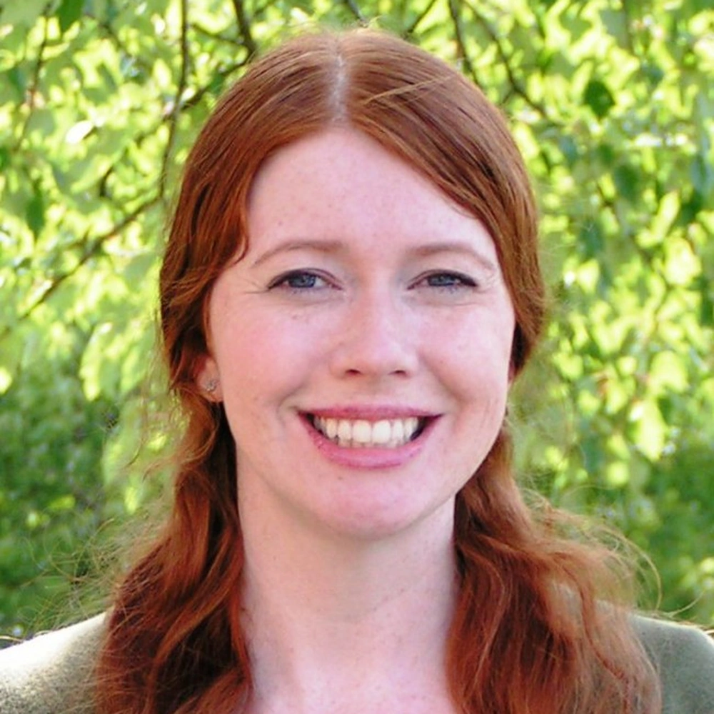 Headshot portrait of Emma Caro, a smiling woman with long auburn hair, photographed outdoors against green foliage.