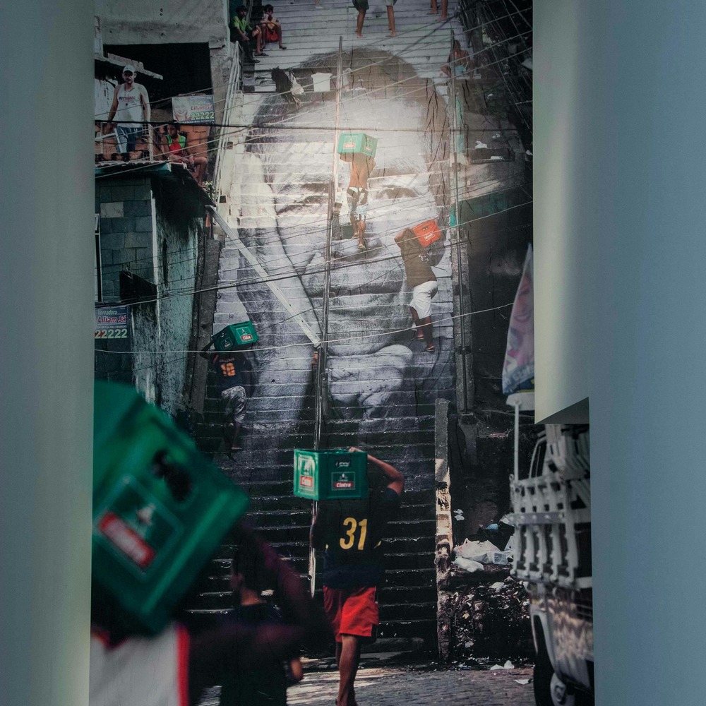 Square detail of JR's exhibition installation at the Contemporary Arts Center: a giant portrait of a face covering a favela staircase mural, with two people carrying green crates.