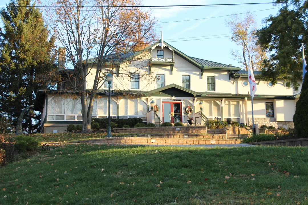 Two-story museum building with white siding and green trim on a grassy hill, red double entrance door with wreaths, small balconies and trees on both sides.