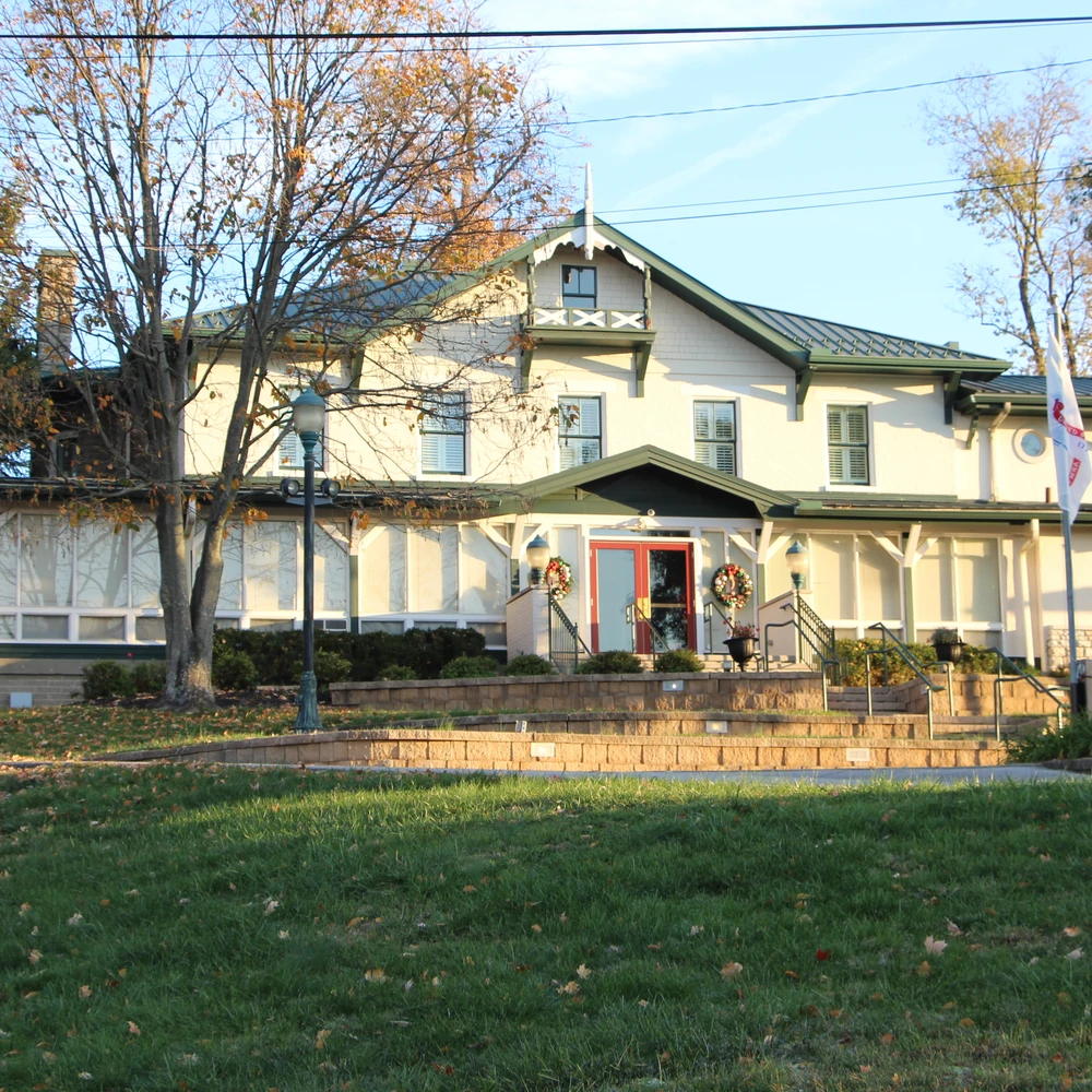 Two-story museum building with white siding and green trim on a grassy hill, red double entrance door with wreaths, small balconies and trees on both sides.