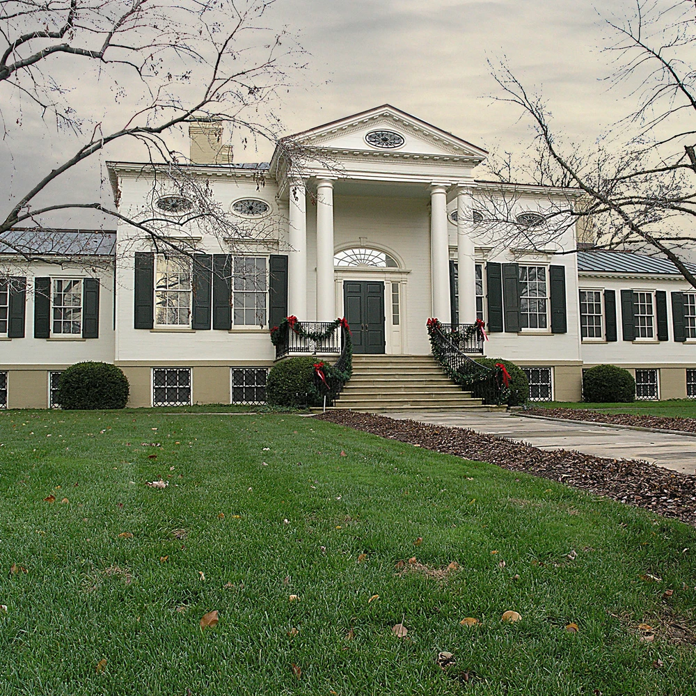 White neoclassical mansion with columned portico and dark green shutters, holiday garlands on the entrance railings, seen across a lawn.