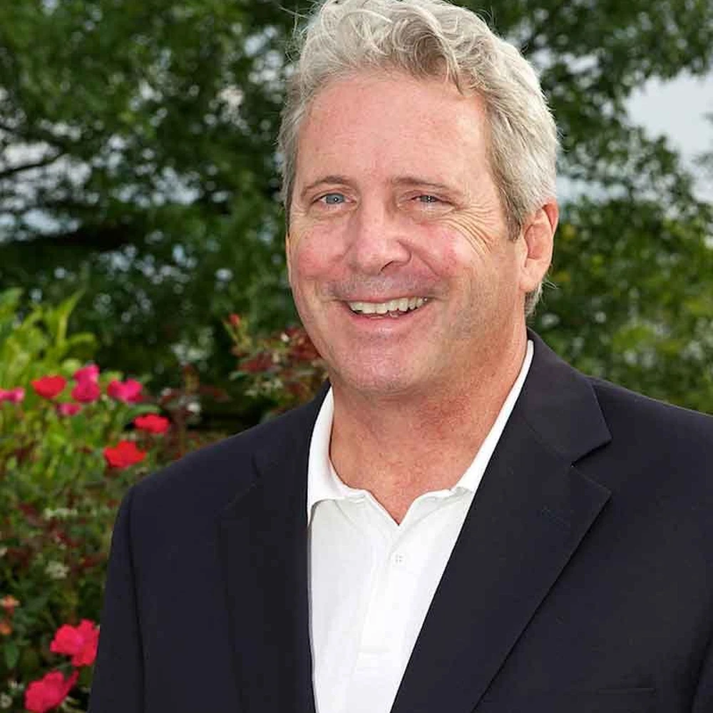 Portrait of Kip Eagen, a smiling man with gray hair wearing a dark blazer, outdoors with red flowers behind him.