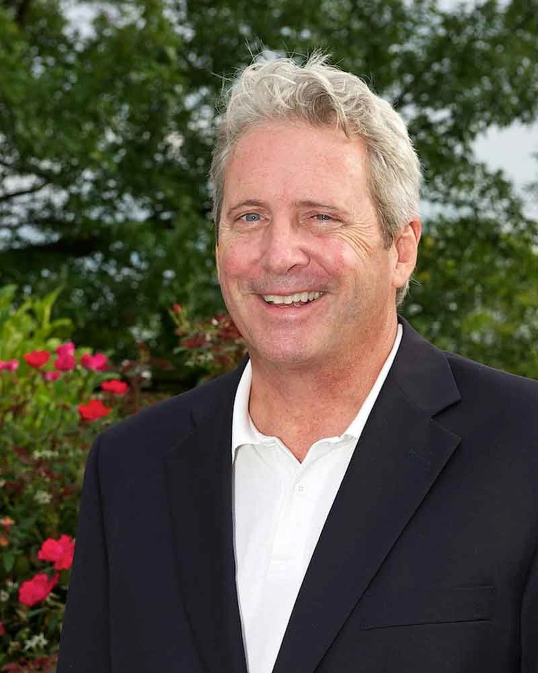 Portrait of Kip Eagen, a smiling man with gray hair wearing a dark blazer, outdoors with red flowers behind him.