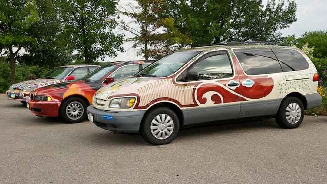 Three painted art cars parked in a row, the nearest a minivan with red swirls.