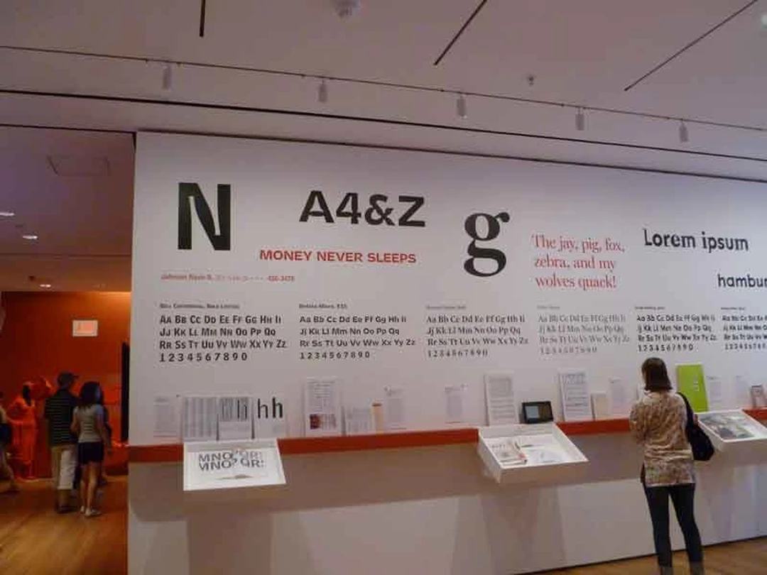 MoMA gallery wall covered with large type specimens and font samples, with visitors looking at them.