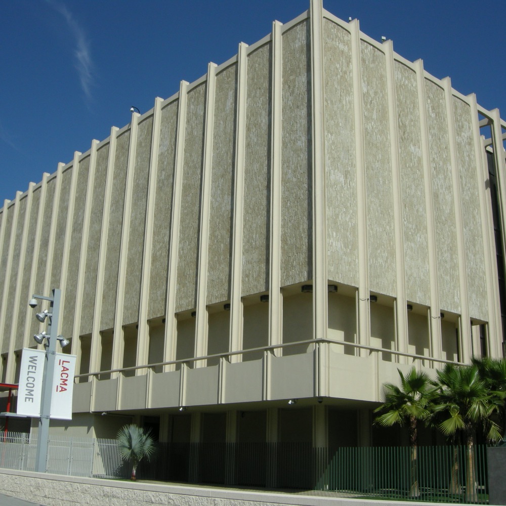 Facade of the Los Angeles County Museum of Art with tall vertical stone fins, WELCOME banners on lampposts, palm trees and a dark statue in front, under a clear blue sky.