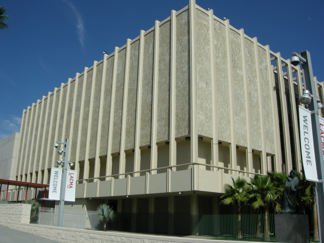 Facade of the Los Angeles County Museum of Art with tall vertical stone fins, WELCOME banners on lampposts, palm trees and a dark statue in front, under a clear blue sky.