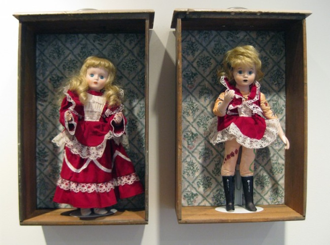 Two dolls in wooden boxes, one in a long red dress with blond curls, the other in a short red dress with tattoos and black boots