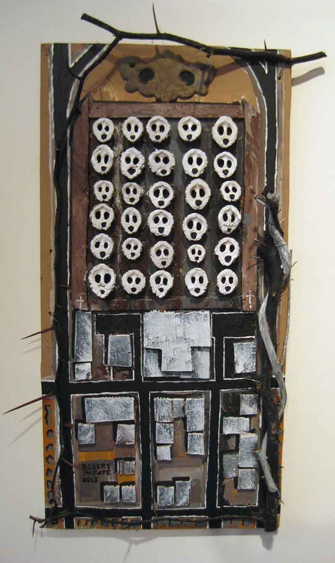 Tall narrow construction with rows of white skull-like faces above a painted city block, signed Robert McFate 2013