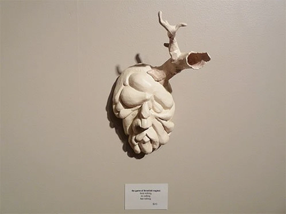 White sculptural mask with an antler-like protrusion mounted on a gallery wall.