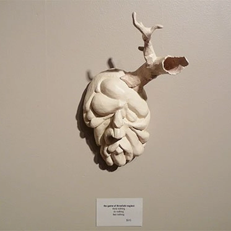 White sculptural mask with an antler-like protrusion mounted on a gallery wall.