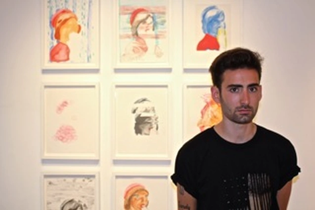Aaron Skolnick, in a black T-shirt, standing in front of his framed drawings at Rare Gallery, New York, 2013.
