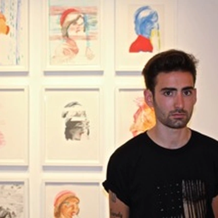 Aaron Skolnick, in a black T-shirt, standing in front of his framed drawings at Rare Gallery, New York, 2013.