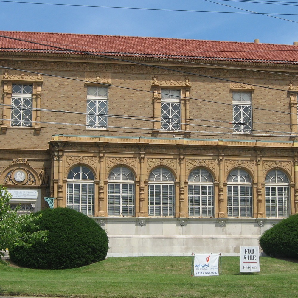 Two-story brick building with a red tile roof and arched windows with ornate stone surrounds, seen across a lawn with bushes; overhead power lines cross the frame.