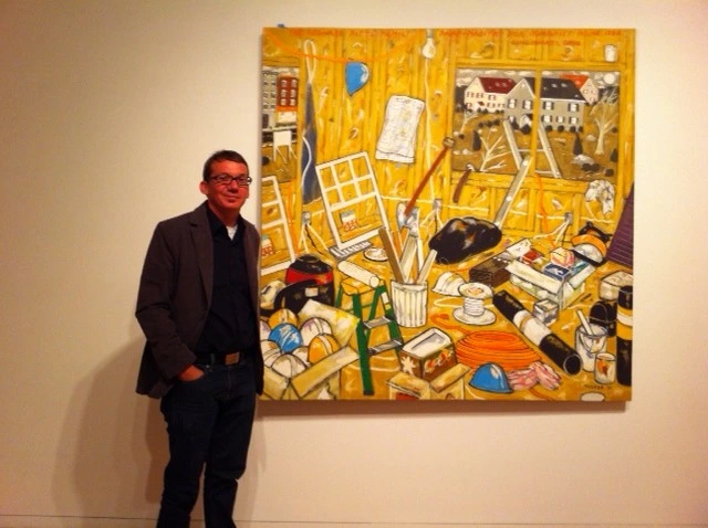 Aaron Cowan in a dark jacket standing beside a large yellow painting of a cluttered studio interior, in a gallery.