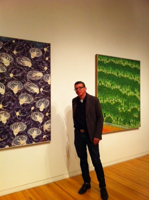 Aaron Cowan standing in a gallery corner between a dark patterned painting and a green painting of grass.