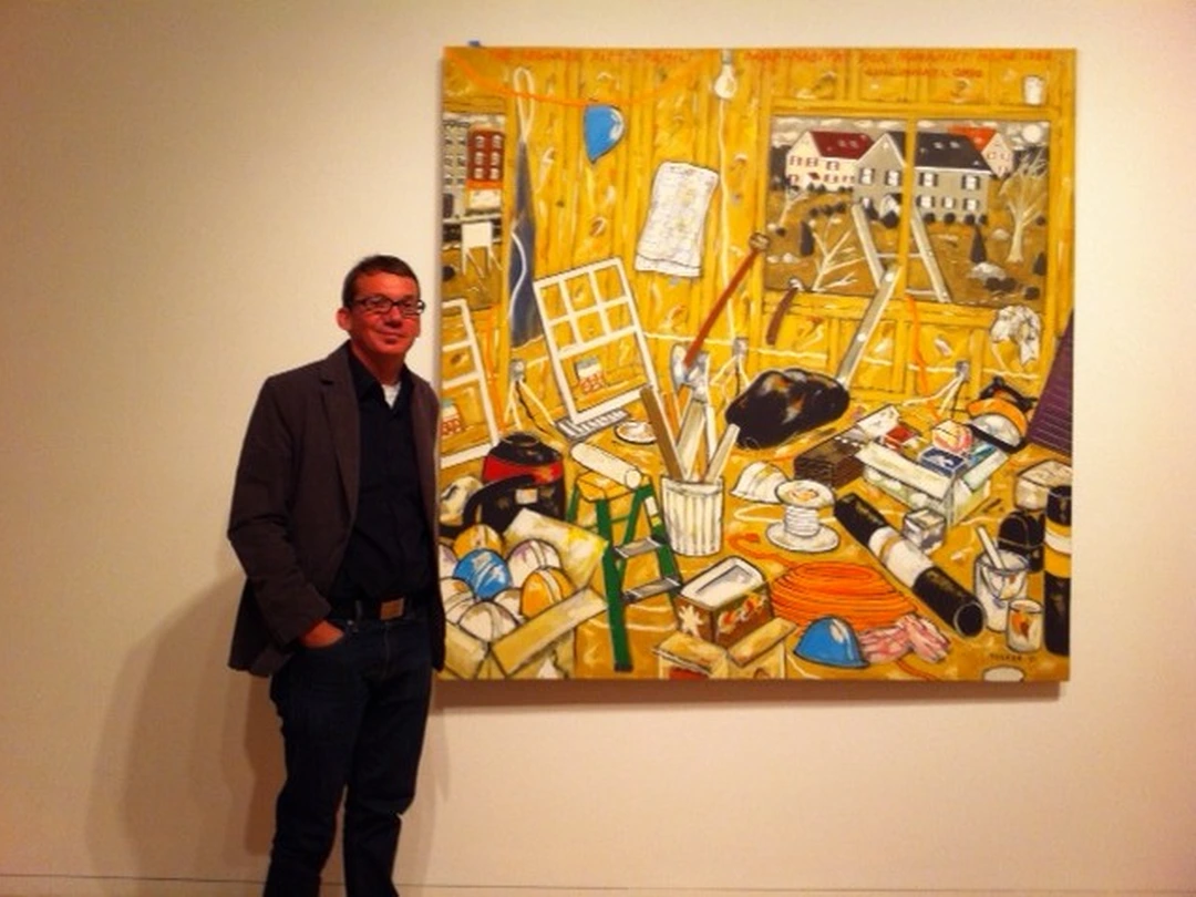 A man in a dark jacket standing beside a large colorful abstract painting in a gallery.