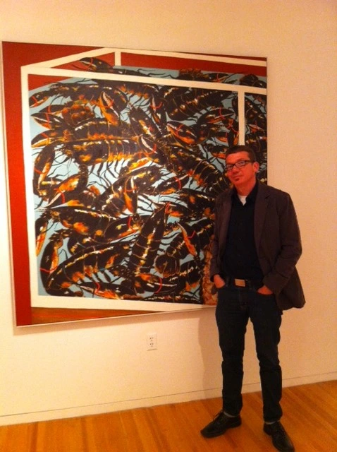 Aaron Cowan standing beside a large painting of orange and black leaf-like forms inside a red frame, in a gallery.