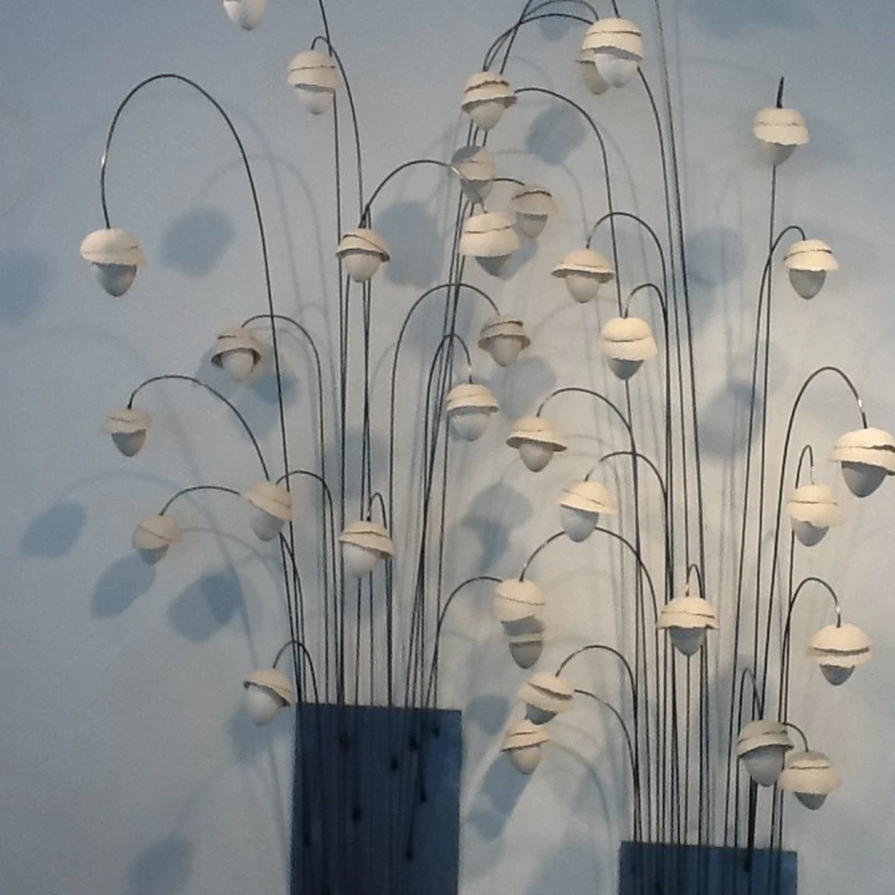 White porcelain blossom forms on slender, arched metal stems rising from a dark rectangular base, against a white gallery wall.