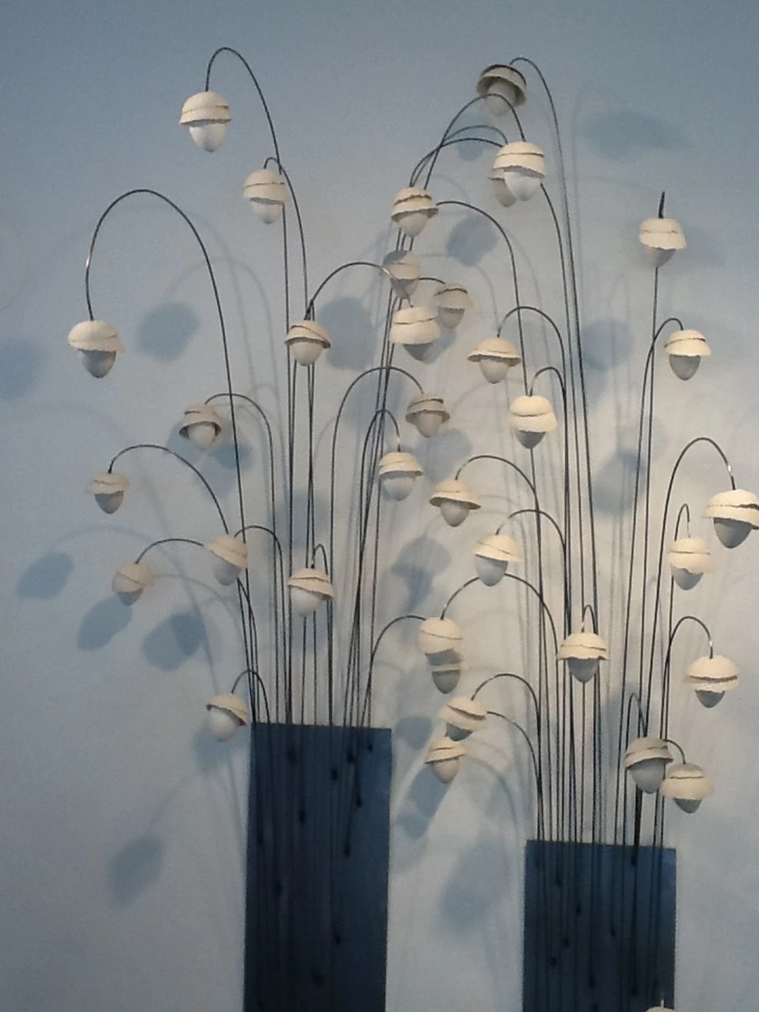 White porcelain blossom forms on slender, arched metal stems rising from a dark rectangular base, against a white gallery wall.