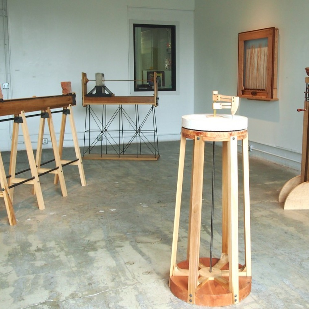 Installation view of the exhibition at Brazee Studios: several wooden kinetic sculptures, including sawhorse structures with bricks, a round table and a tall column.