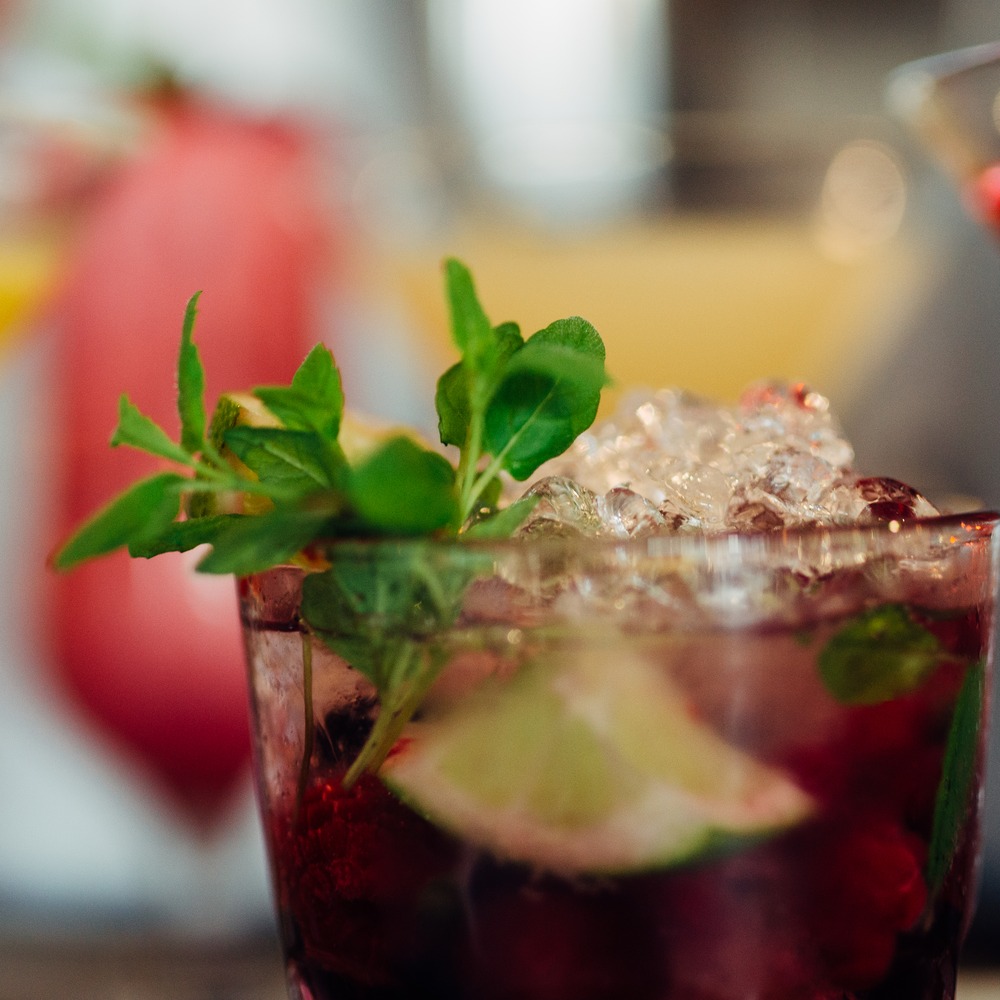Close-up of colorful cocktails on a bar counter: a dark red drink with crushed ice, fresh mint and a lime wedge in focus, with blurred yellow and red cocktails behind.