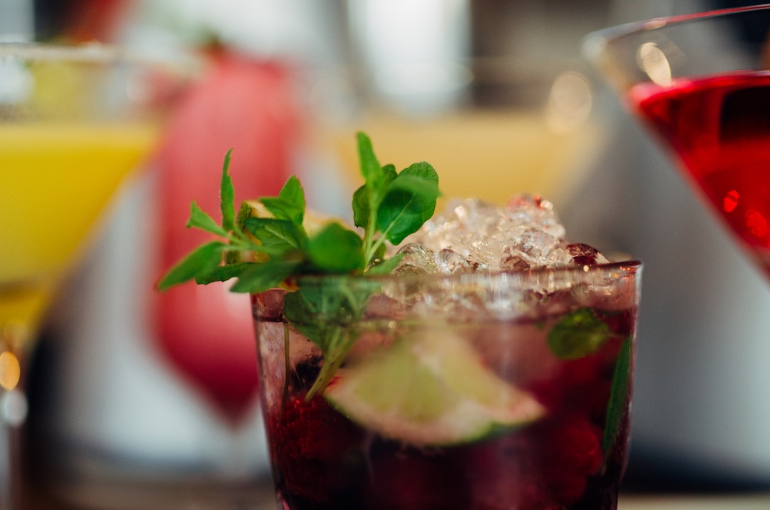 Close-up of colorful cocktails on a bar counter: a dark red drink with crushed ice, fresh mint and a lime wedge in focus, with blurred yellow and red cocktails behind.